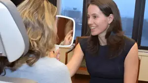 A woman in a dark dress, smiling and holding a mirror for a seated individual with blonde hair, who appears to be having first-time anti-wrinkle injections in a well-lit room with large windows.