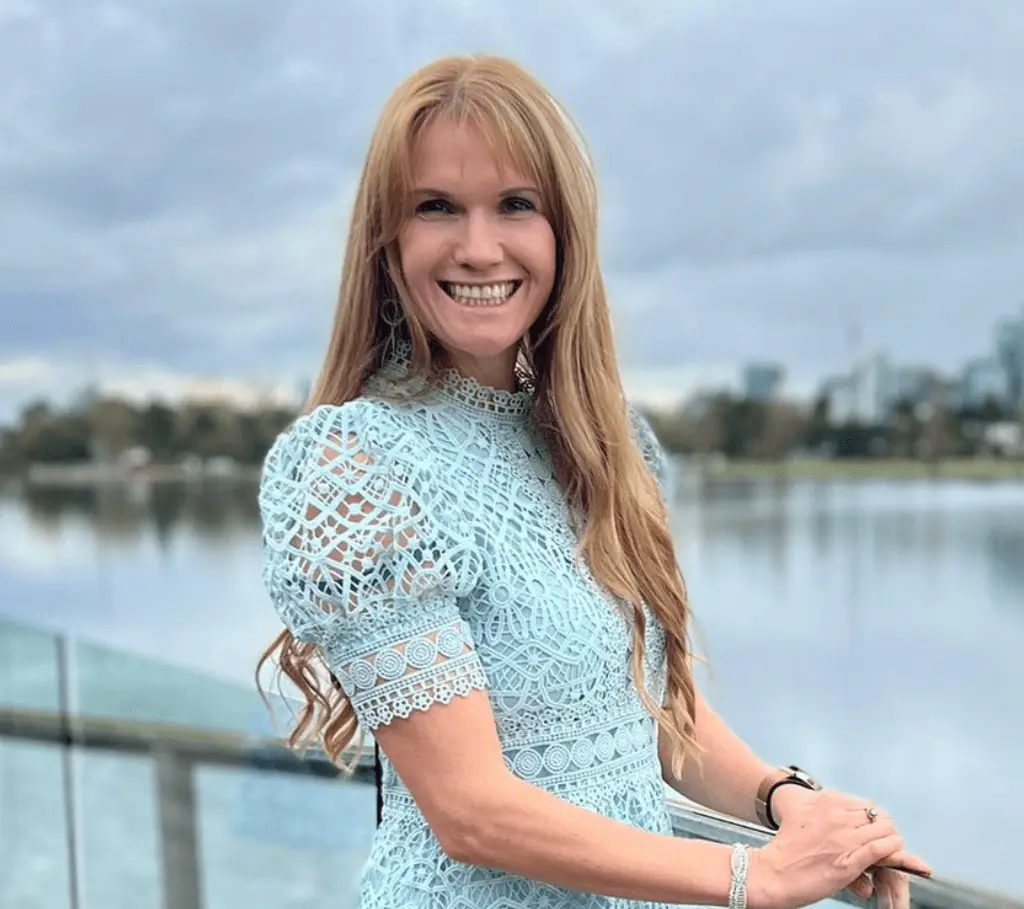 A woman with long, light brown hair smiles while standing by a body of water. She wears a light blue, lace dress and poses with her hands resting on a railing. The background is slightly cloudy, creating an elegant and serene scene perfect for an About page or Description.