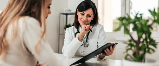 The doctor, holding a clipboard and pen, smiles while discussing GP appointments with a seated female patient in a well-lit office with a plant on the desk.