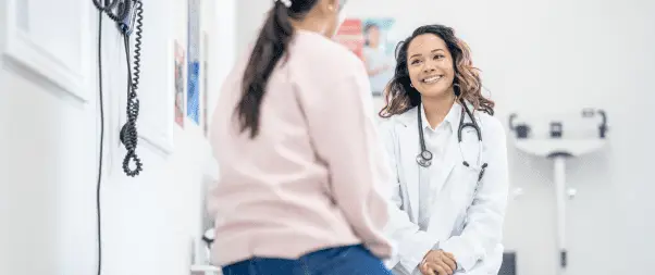 A female doctor in a white coat and stethoscope smiles while discussing Women's Health with a seated patient in a medical office.