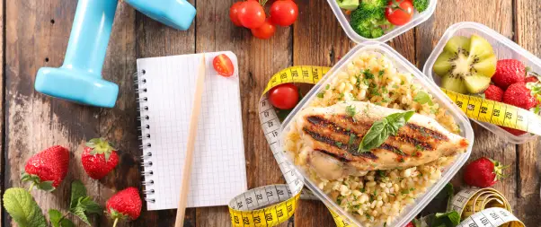 A grilled chicken breast with herbs over grains in a plastic container, surrounded by fruits, vegetables, a notebook, a tape measure, and a dumbbell on a wooden surface—perfect for anyone focusing on their diet and exercise.