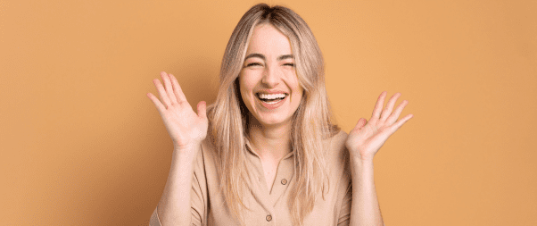 A woman with long blonde hair smiles broadly and holds both hands up in the air in front of an orange background, embodying the joy and confidence often sought through aesthetic treatments.