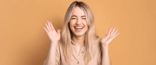 A woman with long blonde hair smiles broadly and holds both hands up in the air in front of an orange background, embodying the joy and confidence often sought through aesthetic treatments.